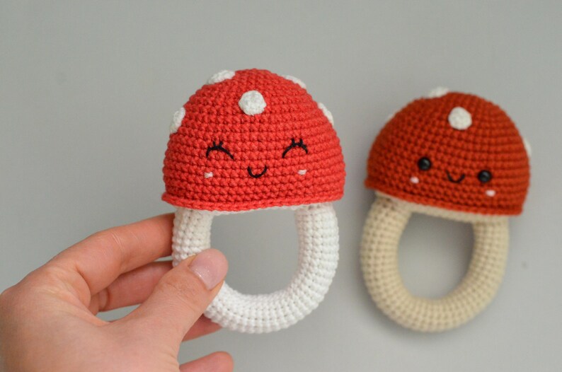 May include: Two crocheted mushroom-shaped baby rattles. One is red with white dots and a smiling face, the other is brown with white dots and a smiling face. Both have white crocheted rings.