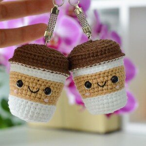 May include: Two crocheted coffee cup keychains with smiling faces. The cups have brown lids, tan bodies, white bottoms, and black eyes. The keychains are held up in front of a blurred background of pink flowers and a white shelf.