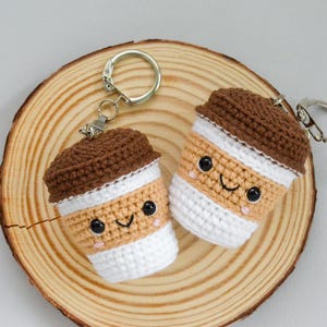 May include: Two handmade crochet coffee cup keychains with smiling faces, brown lids, and white and tan bodies. The keychains are on a wooden slice, perfect for coffee lovers.