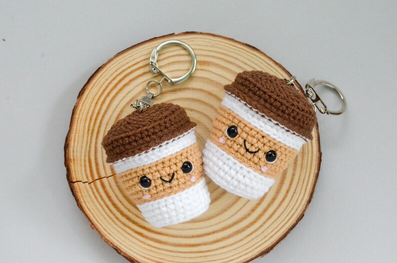 May include: Two handmade crochet coffee cup keychains with smiling faces, brown lids, and white and tan bodies. The keychains are on a wooden slice, perfect for coffee lovers.
