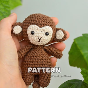 May include: A handmade crochet monkey doll in shades of brown and cream. The monkey has a light cream face, ears, and belly, with black eyes and a small mouth. The word "PATTERN" is visible on the doll. The doll is held in a hand.