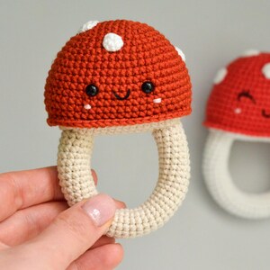 May include: Two crocheted mushroom toys with smiling faces. The mushrooms are red with white spots and have a cream-colored stem. One mushroom is being held by a hand.