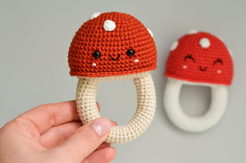 May include: Two crocheted mushroom toys with smiling faces. The mushrooms are red with white spots and have a cream-colored stem. One mushroom is being held by a hand.
