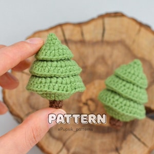 Könnte beinhalten: Zwei kleine grüne gehäkelte Tannenbäume mit braunen Garnstämmen auf einer Holzoberfläche. Der Text "PATTERN @Puppsik_patterns" befindet sich unter den Bäumen.