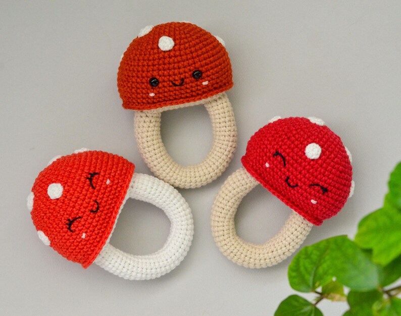 May include: Three crocheted mushroom-shaped teethers with white rings. The mushrooms are red with white polka dots and have embroidered smiling faces.