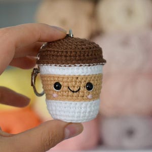 May include: A crocheted coffee cup keychain with a smiling face. The cup is brown, tan, and white, with a silver keyring. The background is blurred, showing other yarn balls.