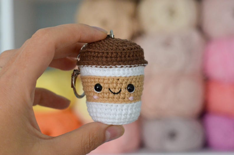 May include: A crocheted coffee cup keychain with a smiling face. The cup is brown, tan, and white, with a silver keyring. The background is blurred, showing other yarn balls.