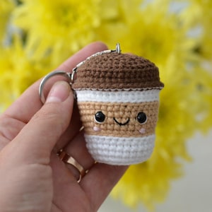 May include: A crocheted coffee cup keychain with a smiling face. The keychain is brown, tan, and white, with a silver key ring. The background is a blur of yellow flowers.