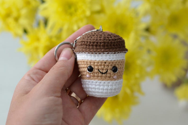 May include: A crocheted coffee cup keychain with a smiling face. The keychain is brown, tan, and white, with a silver key ring. The background is a blur of yellow flowers.