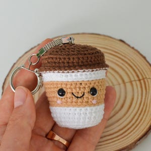 May include: A crocheted coffee cup keychain in brown, tan, white, and pink. The keychain has a smiling face and a brown lid. The keychain is held in a hand, with a silver key ring attached. The background is a wooden slice.
