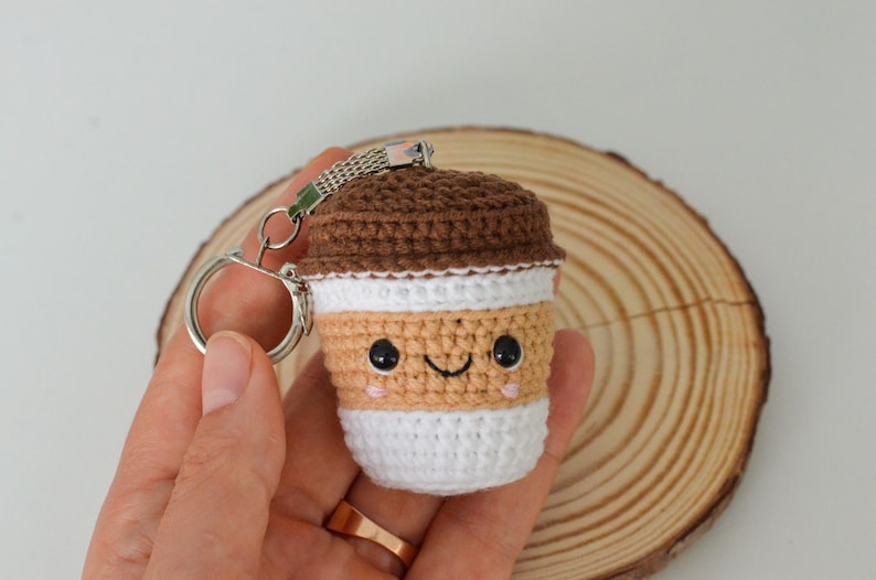 May include: A crocheted coffee cup keychain in brown, tan, white, and pink. The keychain has a smiling face and a brown lid. The keychain is held in a hand, with a silver key ring attached. The background is a wooden slice.