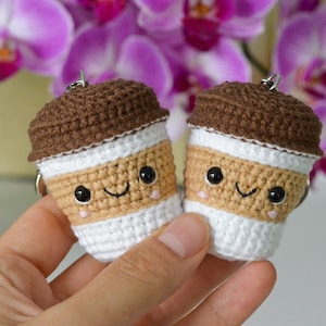 May include: Two crocheted coffee cup keychains. Each keychain features a brown top, tan middle, and white bottom, with a smiling face and black eyes. The keychains are held in front of a blurred background of purple flowers.