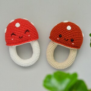 May include: Two crocheted mushroom-shaped baby teethers. The teethers are red and orange with white spots and a smiling face. The teethers are attached to a white crocheted ring.