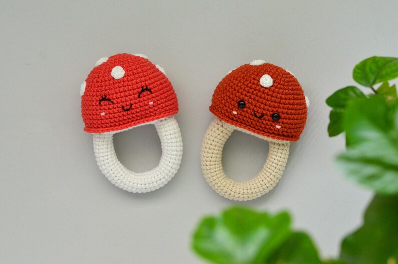 May include: Two crocheted mushroom-shaped baby teethers. The teethers are red and orange with white spots and a smiling face. The teethers are attached to a white crocheted ring.