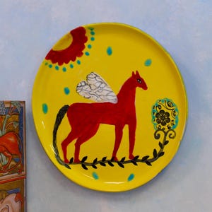 Puede incluir: Un plato de cerámica amarillo con un caballo rojo alado pintado en él. El caballo está de pie sobre una vid negra con hojas verdes. El plato tiene una flor roja con un centro amarillo y puntos azules alrededor.