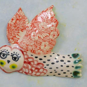 以下が含まれることがあります： 赤と白の花柄の羽を持つセラミック製のフクロウの置物。フクロウの顔は白で、黒い目、黄色のアクセント、赤いディテールがあります。体は白で、黒い斑点と緑色の足があります。