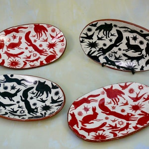 Puede incluir: Cuatro platos ovalados de cerámica con siluetas de animales negras y rojas sobre fondo blanco. Los platos tienen un aspecto rústico, pintados a mano y presentan una variedad de diseños de animales, incluyendo conejos, zorros y pájaros.