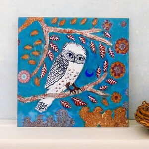 May include: Square artwork depicting a white owl with black patterns, perched on a branch. The background is turquoise, adorned with red, orange, and brown leaves and flowers. A crescent moon is also visible in the artwork.