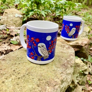 Pode incluir: Duas canecas de cerâmica branca com fundo azul. Apresentam uma ilustração detalhada de uma coruja pousada num galho com flores vermelhas e uma lua cheia. As canecas têm alças e bordas brancas.