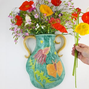 May include: A teal ceramic wall vase with a butterfly and flower design. The vase has two handles and is decorated with yellow stars. A bouquet of red, yellow, and pink flowers is in the vase.
