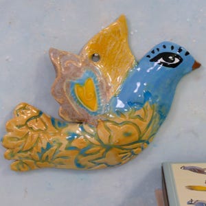 Peut inclure: Un objet décoratif mural en forme d'oiseau en céramique. Le corps est bleu, les ailes jaunes, avec des motifs floraux. L'oiseau a un œil détaillé et un petit trou pour l'accrocher. Les couleurs sont le bleu, le jaune et le marron.