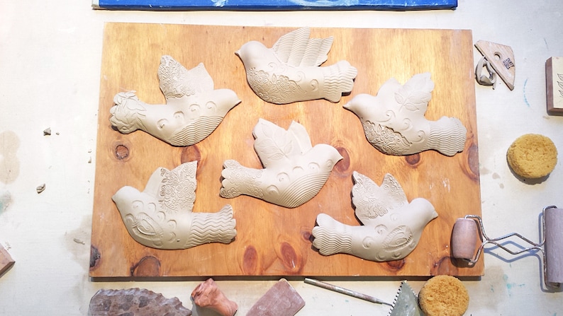 Może przedstawiać: Sześć nieukończonych białych ceramicznych figurek gołębi na drewnianej powierzchni. Gołębie są ułożone w rzędzie, ze skrzydłami rozłożonymi.