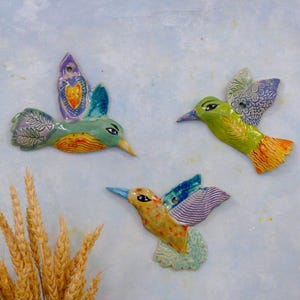 May include: Three ceramic hummingbird wall decorations in flight. The birds are painted in bright colors, including green, orange, blue, and yellow. Each hummingbird has unique wing patterns and details. The background is a light blue.