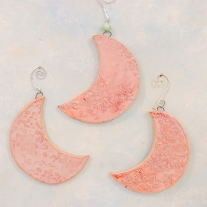 May include: Three pink ceramic crescent moon ornaments with a delicate floral pattern. Each ornament has a silver wire hanger.