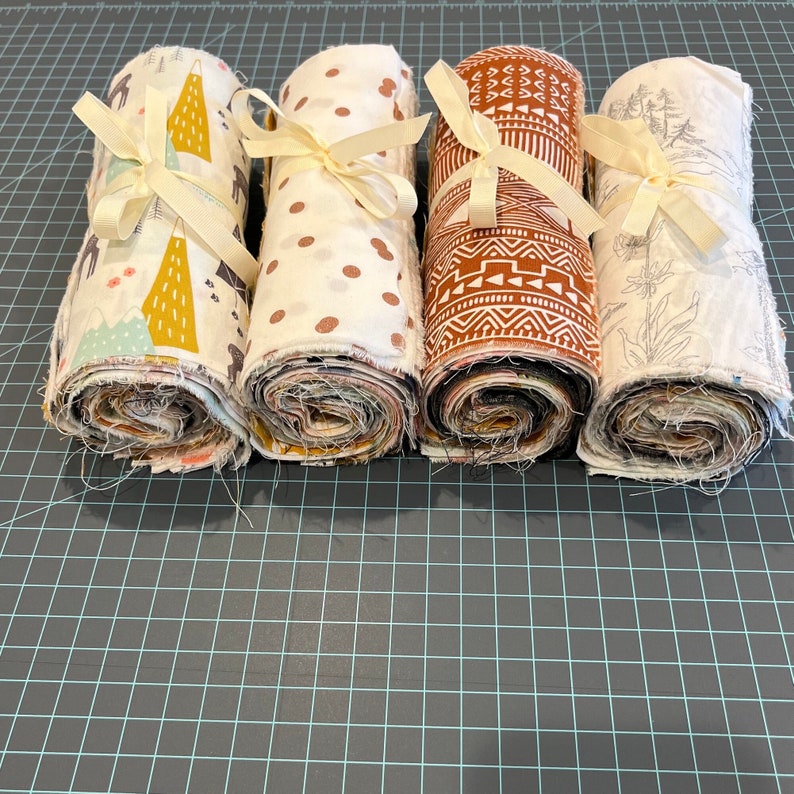 May include: Four fabric bundles rolled and tied with cream-colored ribbons. The bundles feature various patterns: mountains, polka dots, geometric designs, and floral prints. The fabric edges are frayed, and the bundles are arranged on a grid surface.