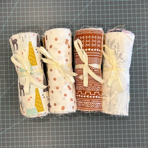 May include: Four rolled fabric bundles tied with cream-colored ribbons. The fabrics feature various patterns: deer and trees, gold polka dots, geometric tribal designs, and a forest scene. The bundles are arranged on a gray grid surface.