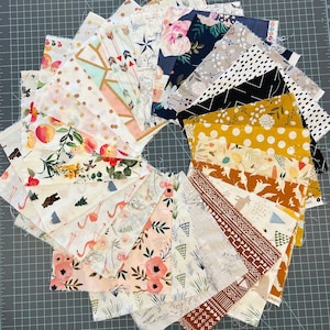 May include: A circular arrangement of fabric squares with various patterns and colors. Designs include florals, arrows, dots, and geometric shapes. The fabrics are primarily white, with accents of pink, blue, gold, and brown. The squares are arranged on a gray cutting mat.