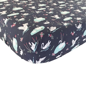 May include: A dark blue fitted crib sheet with a white swan and floral pattern. The sheet features a repeating pattern of swans swimming in a pond with green lily pads and pink flowers.