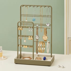 May include: An olive green metal jewelry organizer stand with multiple tiers for hanging necklaces, earrings, and bracelets. The base features two compartments lined with beige fabric, holding rings and a bracelet, while watches hang on the side.