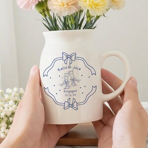 May include: A white ceramic pitcher with a handle, decorated with blue ribbon and text that reads "Bella & Jack Engaged 10.15.25". The pitcher holds a bouquet of pink, peach, and yellow carnations. A small wooden tray with white flowers is in the background.