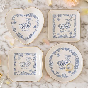 Pode incluir: Quatro pratos de cerâmica brancos para anéis com bordas douradas, cada um com um design floral azul e iniciais dentro de corações ligados. As datas são impressas abaixo dos corações. Os pratos são em forma de coração, quadrados e redondos.