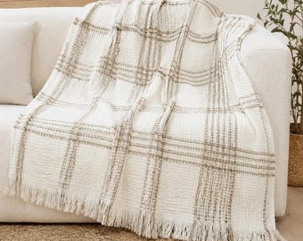 May include: A white waffle-weave throw blanket featuring a subtle beige plaid pattern. The blanket has a textured grid surface and is finished with decorative fringe along the bottom edge, creating a soft and layered appearance across the fabric.