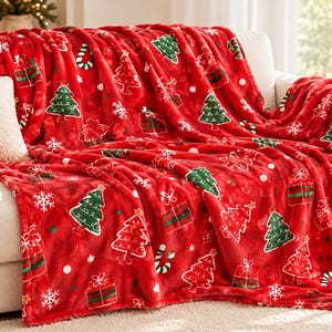 May include: A bright red plush throw blanket featuring a repeating pattern of green Christmas trees, wrapped gift boxes with bows, white snowflakes, and candy canes. The soft fabric has a textured appearance draped over a light-colored sofa.