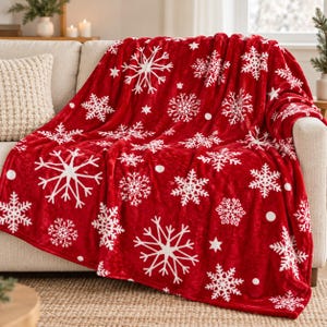 May include: A plush red throw blanket featuring a repeating pattern of white snowflakes and small stars. The fabric has a soft, velvety texture and is draped over a neutral-colored sofa, showcasing the vibrant holiday-themed design.