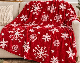 May include: A plush red throw blanket featuring a repeating pattern of white snowflakes and small stars. The fabric has a soft, velvety texture and is draped over a neutral-coloured sofa, showcasing the vibrant holiday-themed design.
