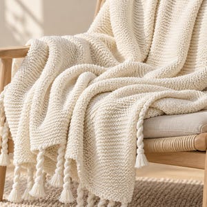 May include: A cream-colored knitted throw blanket with a textured ribbed pattern and thick twisted rope fringe along the bottom edge. The blanket is draped over a light-toned wooden chair with a woven seat, showcasing the soft yarn construction.
