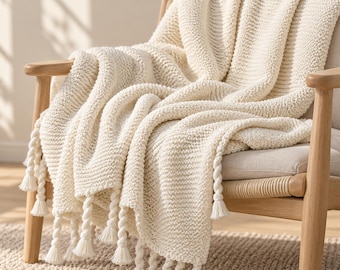 May include: A cream-coloured knitted throw blanket with a textured ribbed pattern and thick twisted rope fringe along the bottom edge. The blanket is draped over a light-toned wooden chair with a woven seat, showcasing the soft yarn construction.