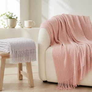 Könnte beinhalten: Eine weiche, hellrosa Strickdecke mit rautenförmiger Textur und langen Fransen, die über einen weißen Sessel drapiert ist, sowie eine gefaltete hellgraue Version derselben Decke auf einem hölzernen Hocker.