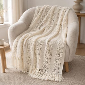 May include: A cream-coloured knit throw blanket draped over a textured armchair. The blanket features alternating vertical stripes of solid knit fabric and an openwork lace pattern, finished with long, knotted fringe along the bottom edge.