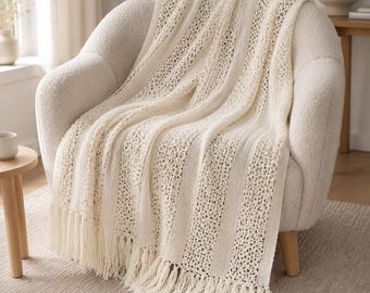 May include: A cream-coloured knit throw blanket draped over a textured armchair. The blanket features alternating vertical stripes of solid knit fabric and an openwork lace pattern, finished with long, knotted fringe along the bottom edge.