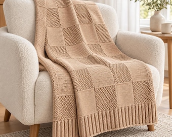 May include: A light tan knitted throw blanket featuring a checkered pattern of alternating smooth stockinette and textured seed stitch squares. The blanket has a wide ribbed border along the bottom edge and is draped over a cream-coloured armchair.