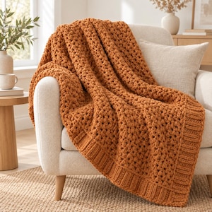 Könnte beinhalten: Eine grob gestrickte Wohndecke in einem kräftigen gebrannten Orange mit einem strukturierten Noppenmuster in der Mitte und einem gerippten Rand, die über einen hellbeigen Polstersessel mit einem passenden Kissen drapiert ist.
