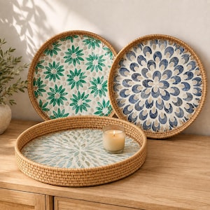 May include: Three round woven rattan trays with raised edges. One tray features a green floral pattern, another has a blue petal design, and the third displays a light green sunburst motif. A small glass candle holder sits in the center tray.