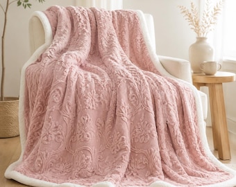 May include: A plush, dusty rose throw blanket featuring a raised, textured damask floral pattern across the surface. The blanket is finished with a thick, soft white sherpa-style trim along all edges, providing a distinct contrast in colour and texture.