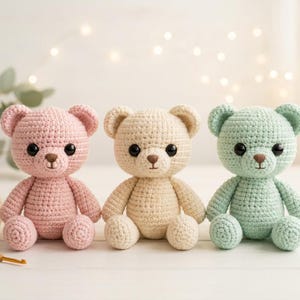 May include: Three handmade crochet teddy bears in pastel colors: pink, cream, and mint green. Each bear has black safety eyes and a brown nose. A ball of yarn and a crochet hook are in the background.