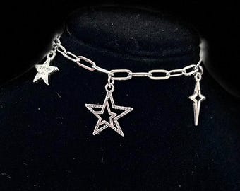 Może przedstawiać: Srebrny naszyjnik typu choker z trzema zawieszkami w kształcie gwiazd. Łańcuszek składa się z połączonych ogniw. Jedna gwiazda to pełny kontur, druga to gwiazda w gwieździe, a trzecia to spiczasta gwiazda.